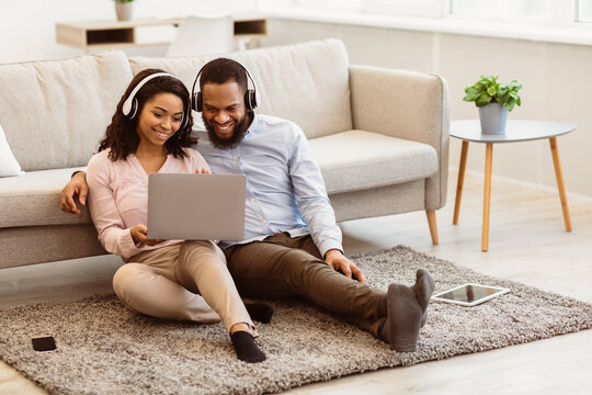 Virtual Communication. Happy black couple in headphones having online video conference with friends or family using computer, waving hand to web camera, sitting on the floor carpet in bedroom at home
