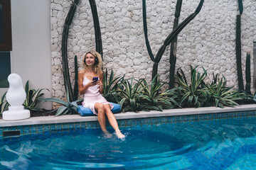 Young woman relaxes by pool, enjoying music in headphones while browsing smartphone—illustrating modern tech integration in leisure and self-care rituals.