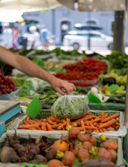 Customer hand selecting fresh vegetables at outdoor market stall, displaying colorful carrots, beets, and leafy greens with blurred background and copy space