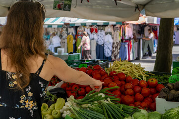 Woman choosing fresh produce at a vibrant outdoor market with colorful stalls and natural lighting on a sunny day.