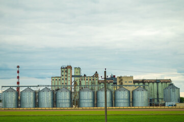 Grain silos and industrial buildings stand near a field under cloudy sky, modern agriculture and production © kazakova0684