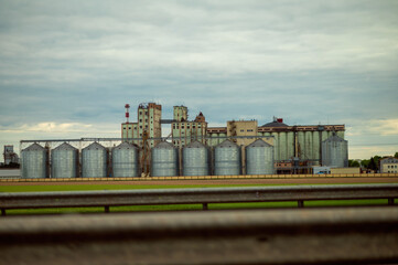 Grain silos and industrial buildings stand near a field under cloudy sky, modern agriculture and production © kazakova0684