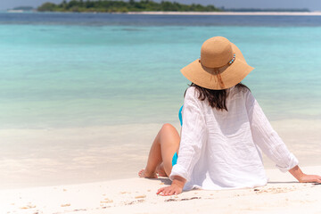 back view woman relax on summer resort, looking at the ocean wearing straw hat and white shirt sits on white sandy beach , peaceful tropical moment in maldives, summer relaxation, travel escape.