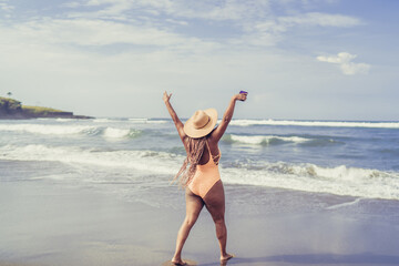 Woman with braids raises arms holding smartphone, celebrating personal freedom, digital independence and joy through expressive mobile technology on a tropical shoreline.