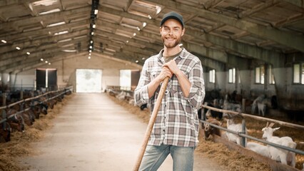 Zooming in handsome Caucasian farmer standing in barn while goats eating hay. Attractive male with beard wearing boots, work uniform and holding pitchfork. Joyfully smiling at camera. © VAKSMANV