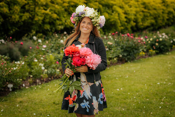 Smiling woman in flower crown holding vibrant peony bouquet, standing in a blooming garden during a...