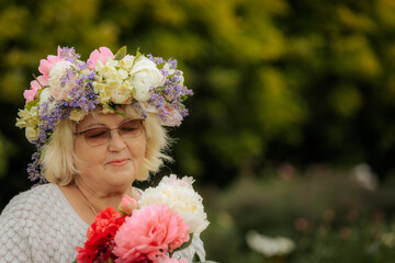 Naklejka premium Elderly woman smiling in a garden, wearing a colorful floral crown and holding a vibrant bouquet of peonies and other flowers.
