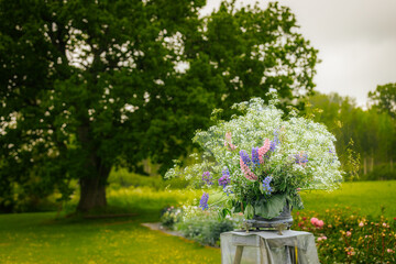 A vibrant bouquet of wildflowers in white, purple, and pink set on a stand in a lush garden, with a...