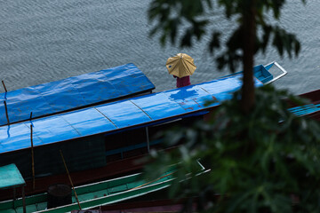 Asiatische Langboote mit blauen Dächern am Fluss bei Regen, eine stehende Person unter gelbem...