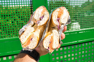 Hand is holding two halves of a fresh whole wheat bagel with smoked salmon, cream cheese, red onions and cucumber, wrapped in aluminium foil, in front of a green metallic fence