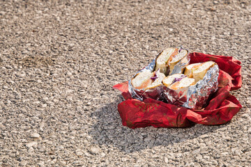 Halves of bagels wrapped in aluminum foil and placed on a red napkin sit on an asphalt surface,...