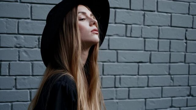 Beautiful young mysterious blond girl in black hat and black jacket on gray brick background