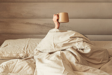 A hand holding a coffee mug emerging from a cozy bed.