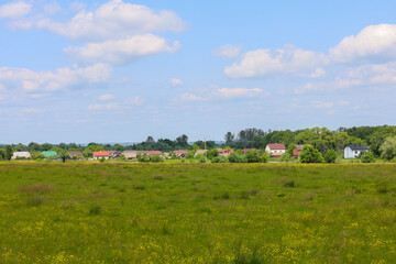 A rural landscape with a large green field, behind which village houses and a dense forest belt are...