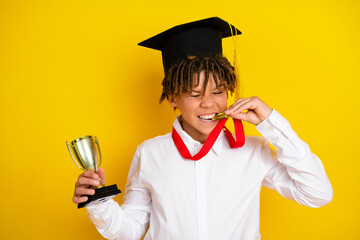 Joyful schoolboy celebrating success with trophy and medal in academic attire on vibrant yellow...