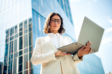 Confident young businesswoman using a laptop outdoors near modern glass buildings, representing...