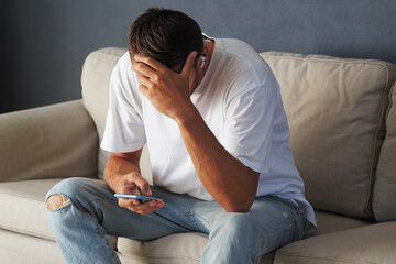 Man struggling, while scrolling social media on smartphone, sitting on sofa at home. 