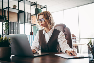 Confident businesswoman in professional attire working in an office space at a desk with a laptop and documents © deagreez