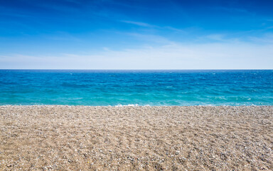 Pebble beach stretches along calm turquoise sea under bright blue sky. Sea and beach meet at clear horizon on sunny summer day. Peaceful coastal landscape without people or buildings