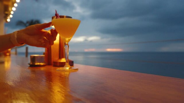 Woman Placing Orchid Flower into Yellow Cocktail in Martini Glass on Bar Counter at Oceanfront Sunset as Camera Pulls Back, Tropical Drink Concept