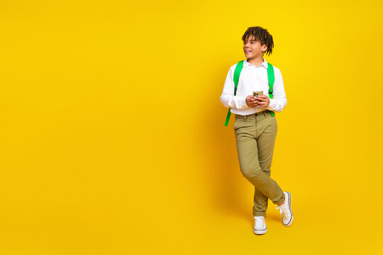 Cheerful young schoolboy in stylish uniform enjoying time with smartphone while standing against vibrant yellow background