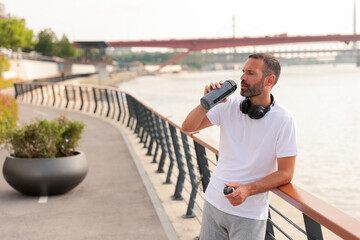 Man enjoying water break while jogging near a river in a sunny afternoon by the waterfront promenade