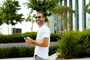 Smiling man in sunglasses enjoys leisurely stroll while texting in sunny urban park setting