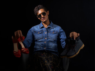 Homme assis avec rouge à lèvre et lunette de soleil qui tient ne paire de chaussure masculine et une paire de chaussure très féminine à talons