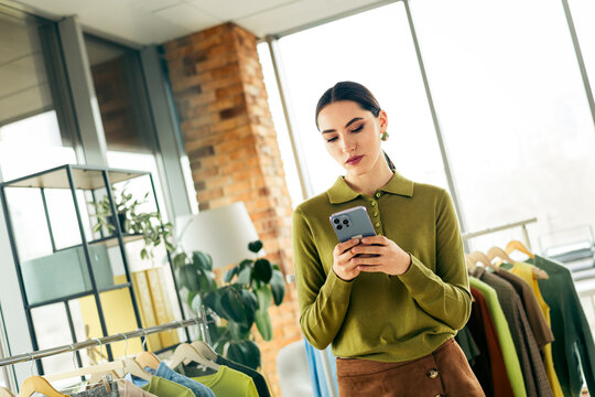 Starting a small business female tailor checks mobile in modern atelier surrounded by racks of clothing