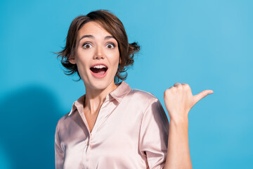 Young woman in pink satin blouse pointing her thumb back with excitement against blue background