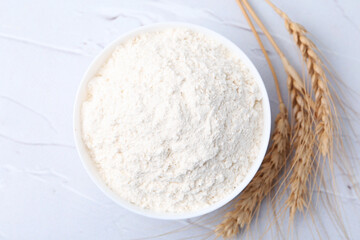 Wheat flour in bowl and spikes on white table, flat lay