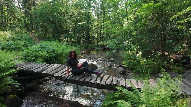 A person finds joy and tranquility on a rustic bridge over a babbling stream, surrounded by vibrant greenery in a serene forest, inviting deep reflection and relaxation in natures beauty