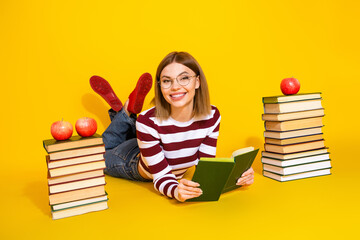 Smiling young woman reading a book, surrounded by stacks of book with apples, on a striking yellow...