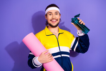 Happy man in retro sportswear holding a yoga mat and water bottle on a vibrant purple background,...