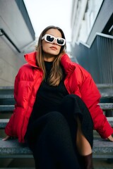 Fototapeta premium Stylish woman in red puffer jacket sitting on steps. She is wearing sunglasses and looking up