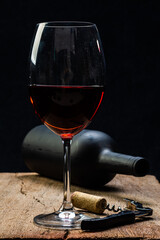 
Glass of red wine on the rustic wooden table, with corkscrew beside it and defocused wine bottle on black background.