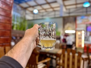 A hand joyfully holds a beer mug in a lively bar with friends and family around. A hand...