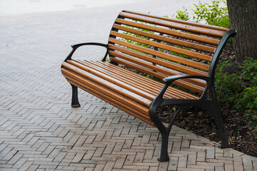 A sturdy and durable wooden park bench sits invitingly along a brick sidewalk right next to a large...