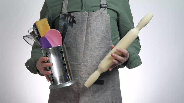 A man in a kitchen apron holds in his hands the inventory for cooking a dish, a rolling pin for rolling out dough. A man is an amateur cook with different inventory, which makes cooking easier and mor