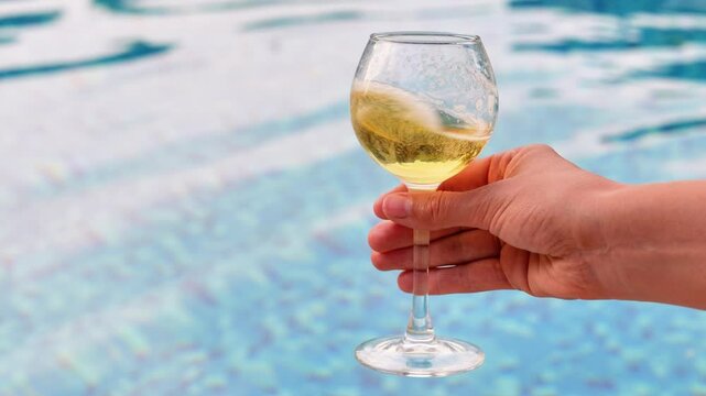 A hand holds a glass of white wine with a swimming pool blurred in the background, suggesting a relaxed, summery setting.