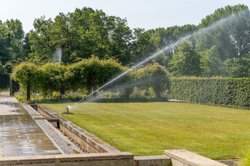 An automatic lawn sprinkler waters a well-kept garden in the sunshine. Hedges and pergolas with...