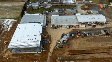 Workers and heavy machinery are busy at a construction site where new commercial buildings are emerging. The area is surrounded by houses and fields, indicating an active development project.