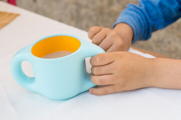 A boy in blue pajamas at breakfast holds a two-handled cup filled with vegetable breakfast drink