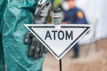 Person in protective gear, sandy area, greenery backdrop, full-body suit, gloves, ATOM sign,...