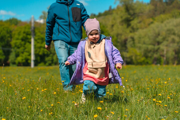 Fototapeta premium A little child girl in a hat runs through a spring field with dandelions and grass