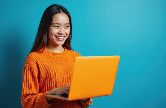 Young woman using laptop computer creating project on blue background. Smiling girl works online with pleasure, wearing orange sweater. Freelancer, student works, study, browsing, lifestyle, winter,