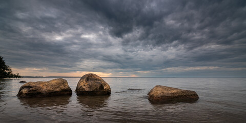Evening lake landscape. Large boulders jut out of calm water near the shore of a large reservoir under a dramatic cloudy sky. Widescreen side view in 10x5 format