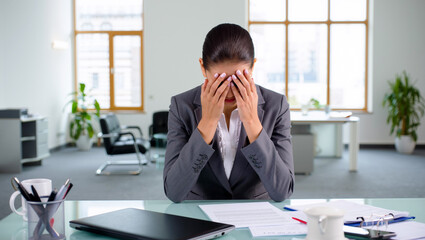 A woman in a business suit holds her head in her hands, displaying signs of stress and depression at her desk in a bright office space filled with plants and furniture. Burnout concept. Generative AI.