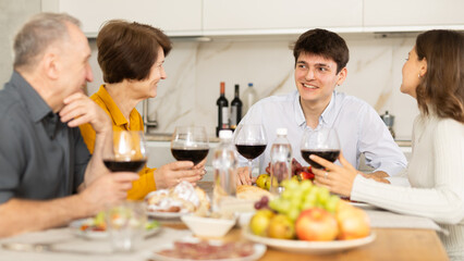 Carefree young guy enjoying family dinner with wife and friendly aged parents-in-law, sharing stories and laughing over wine and appetizers in cozy kitchen