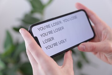 Cyber bullying. Woman holding smartphone with insults on blurred background, closeup